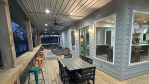 Una terraza techada con piso de madera, una mesa de comedor con sillas y un espacio de estar con taburetes.