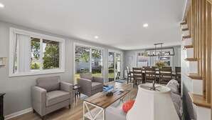 A spacious living area with a sofa, armchairs, a coffee table, and a dining table with chairs. Large windows provide natural light and views of the outdoors. - 7-bedroom home that sleeps 14.  5-minute walk to Moody Beach (Wells)