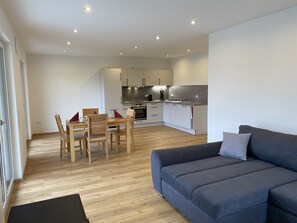 A modern living room with a grey sofa, wooden dining table, and chairs. A kitchen with white cabinets and appliances is visible in the background. - Ferienwohnung in Ravensburg im Grünen, Neubau, Natur (Ravensburg)