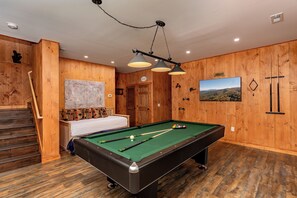 Une table de billard avec queues et billes, un lit avec des coussins à motifs, un escalier et des murs en bois ornés d’un tableau.