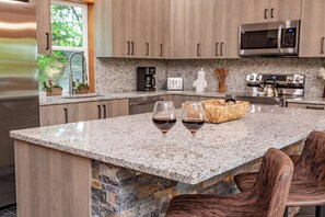 A kitchen with granite countertops, a stone backsplash, wooden cabinets, a built-in microwave, a coffee maker, and a toaster. There are two wine glasses on the counter.