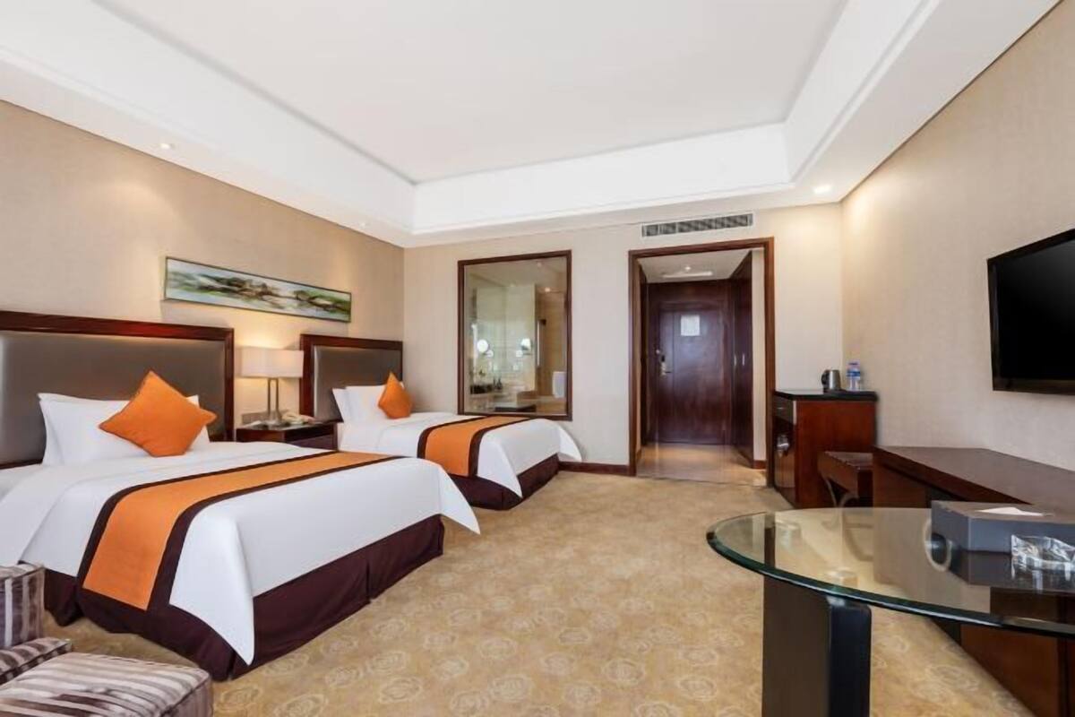 Quarto de hotel com duas camas, uma mesa de vidro, televisão e porta para um corredor.