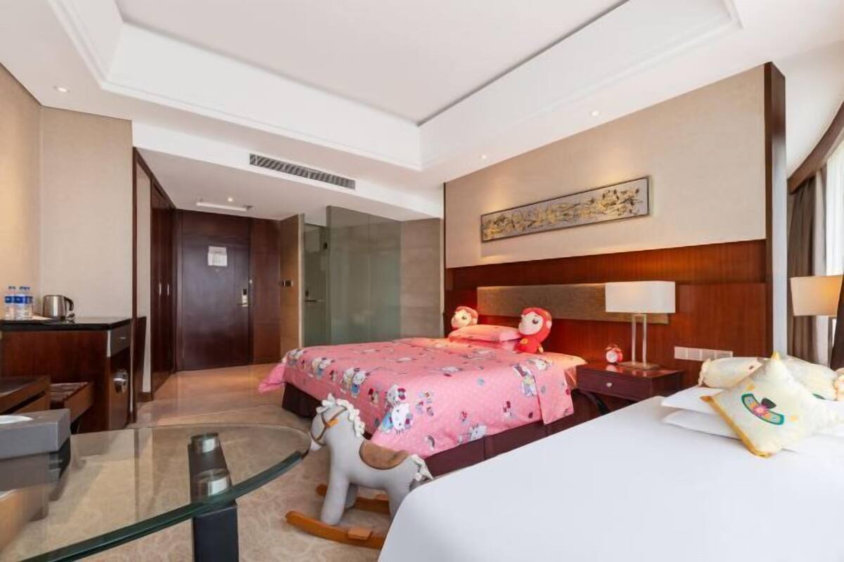 Quarto de hotel com uma cama, uma escrivaninha, uma mesa de vidro, um cavalinho de baloiço e uma mesa de cabeceira com uma lâmpada.