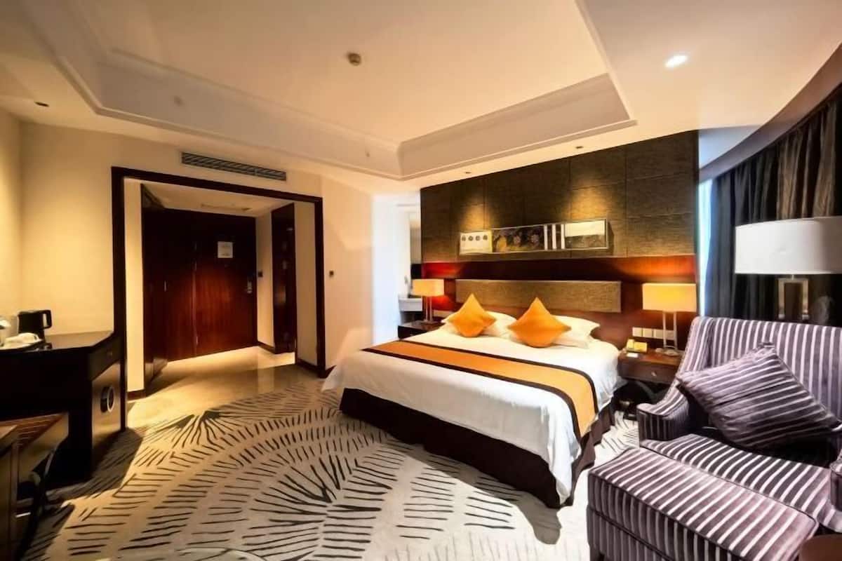 Quarto de hotel com uma cama grande, uma poltrona listrada, uma escrivaninha e um tapete estampado.