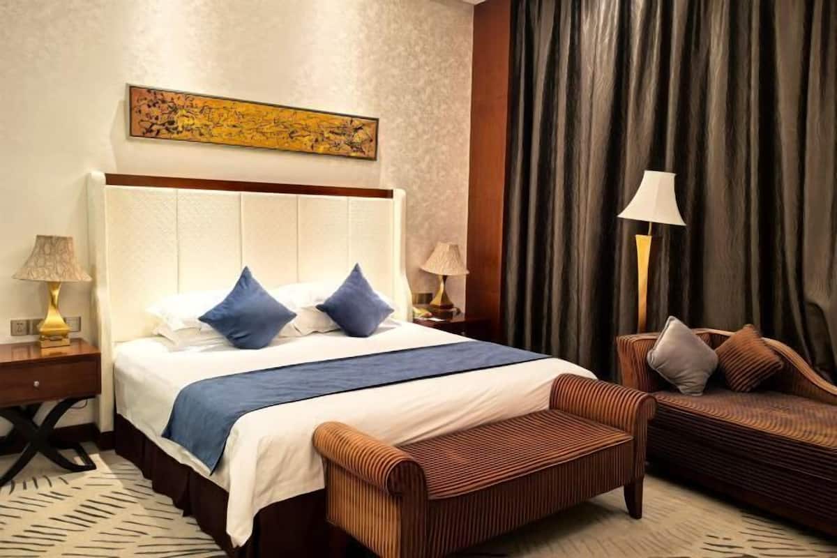 Quarto de hotel com uma cama grande, duas mesas de cabeceira, uma cadeira, um sofá, um quadro na parede e uma cortina.