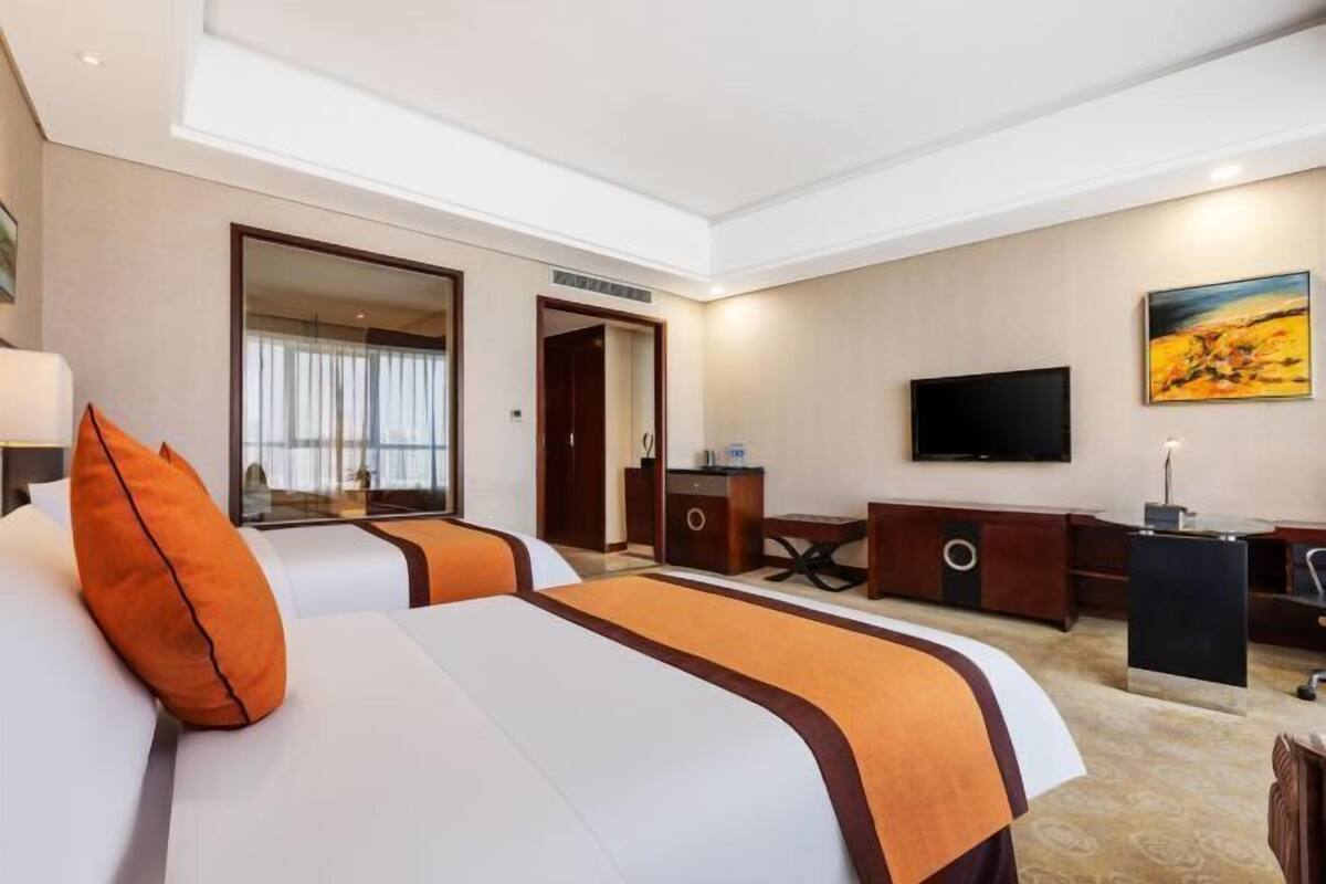Quarto de hotel com duas camas, uma escrivaninha, uma televisão e um quadro na parede.