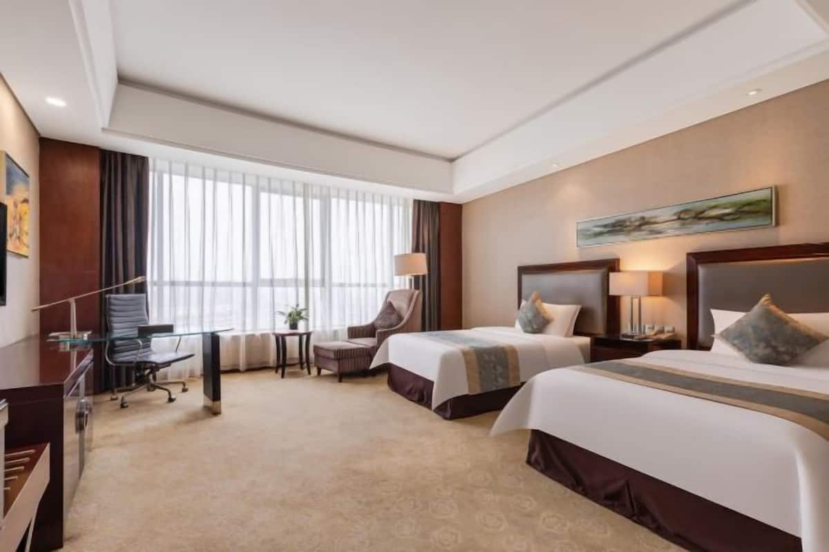 Quarto de hotel com duas camas, uma mesa, uma cadeira e uma janela grande com cortinas.