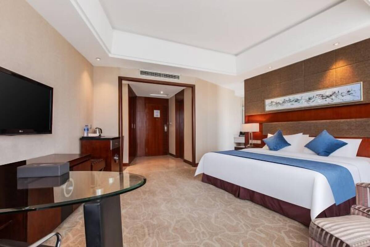 Quarto de hotel com cama, escrivaninha, televisão e um quadro na parede.