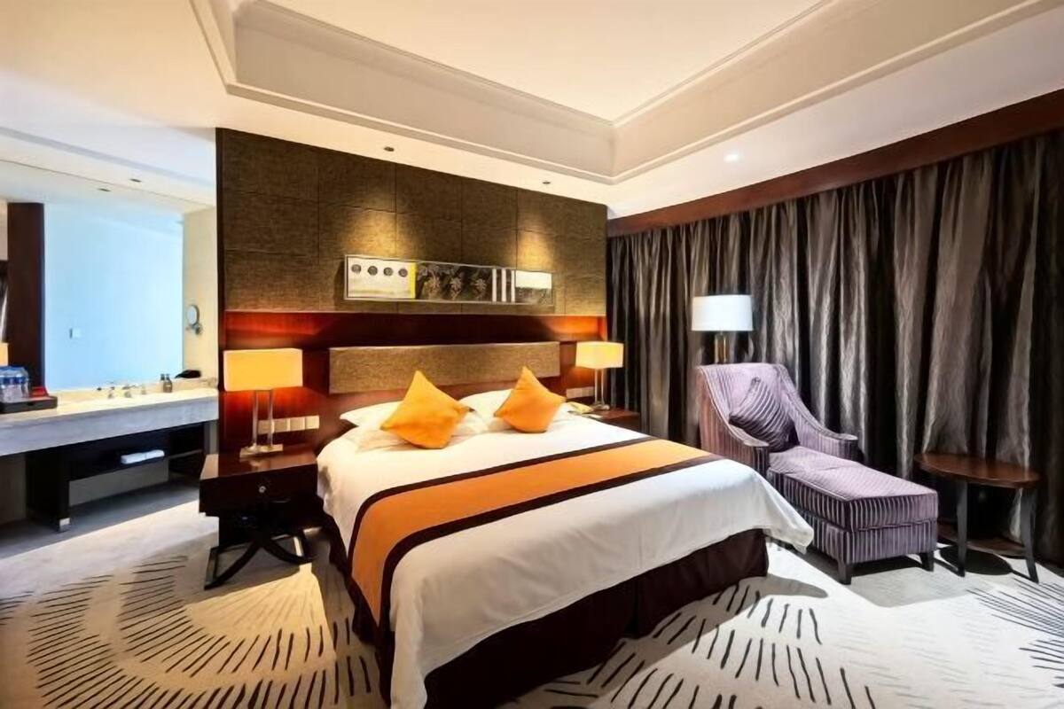 Quarto de hotel com uma cama grande, uma escrivaninha, uma cadeira, um espelho e uma janela com cortinas.