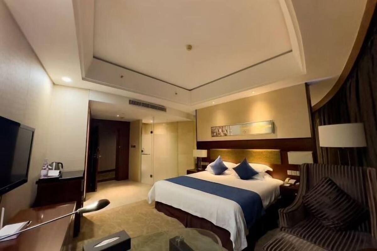 Quarto de hotel com cama, mesas de cabeceira, escrivaninha, cadeira, televisão e um quadro na parede.