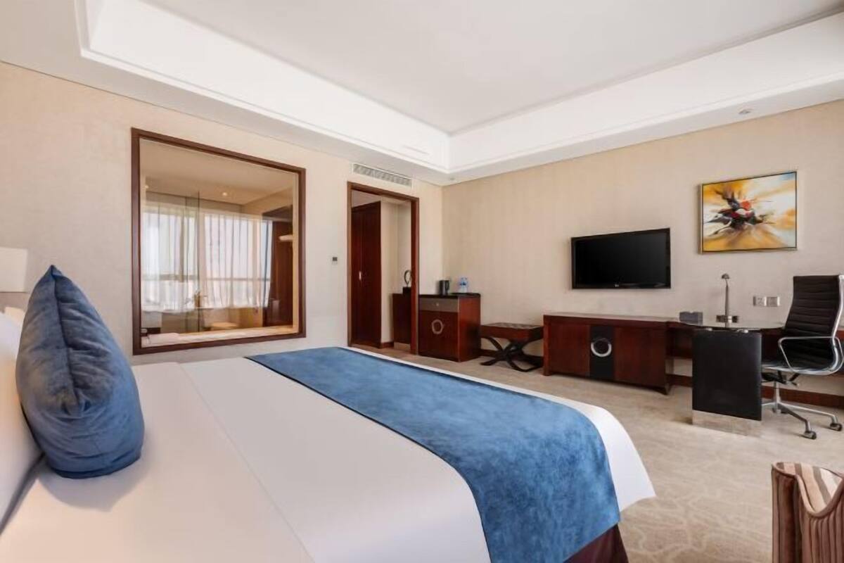 Quarto de hotel com cama, escrivaninha, cadeira, televisão e um espelho grande.