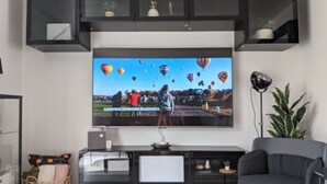 A modern living room with a large flat-screen TV mounted on the wall, a grey sofa, a small side table, and a wicker basket with cushions.