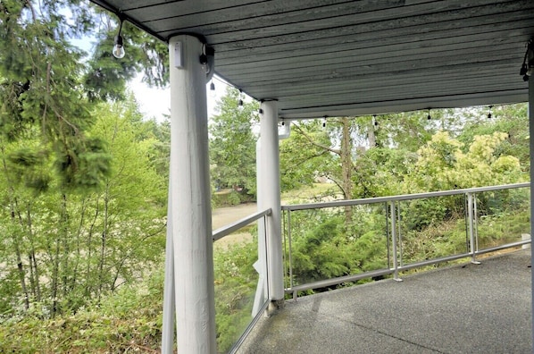 A covered outdoor area with a metal railing, surrounded by trees and greenery. - Heron's Landing - 2BDRM - Oceanfront (Nanoose Bay)