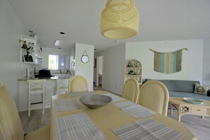 A dining area with a table set for four, a kitchen in the background, and a living area with a sofa and decorative items.