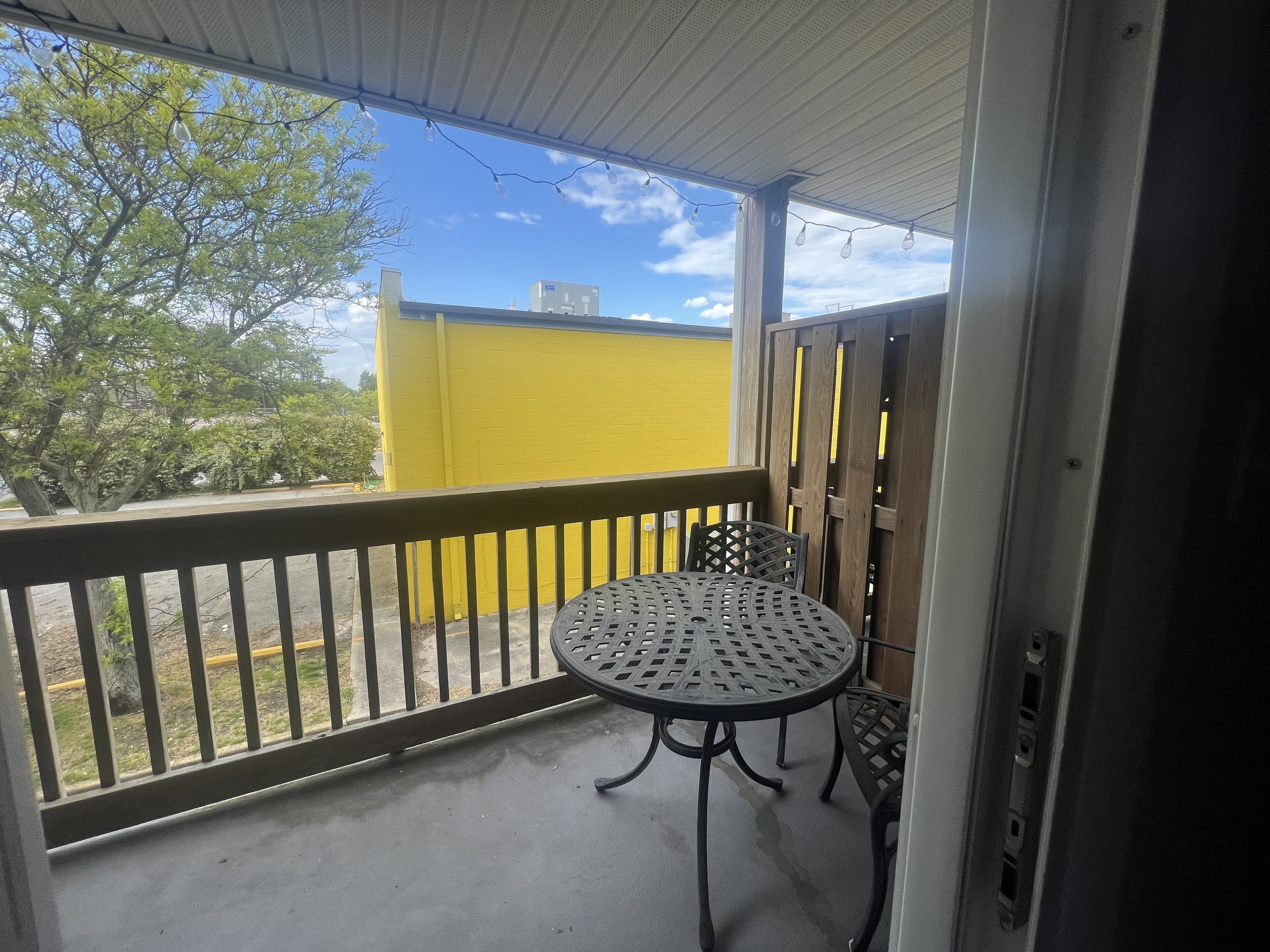 A covered balcony with a metal table and chairs, a yellow wall in the background, and a tree outside.