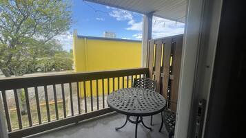 A covered balcony with a metal table and chairs, a yellow wall in the background, and a tree outside.