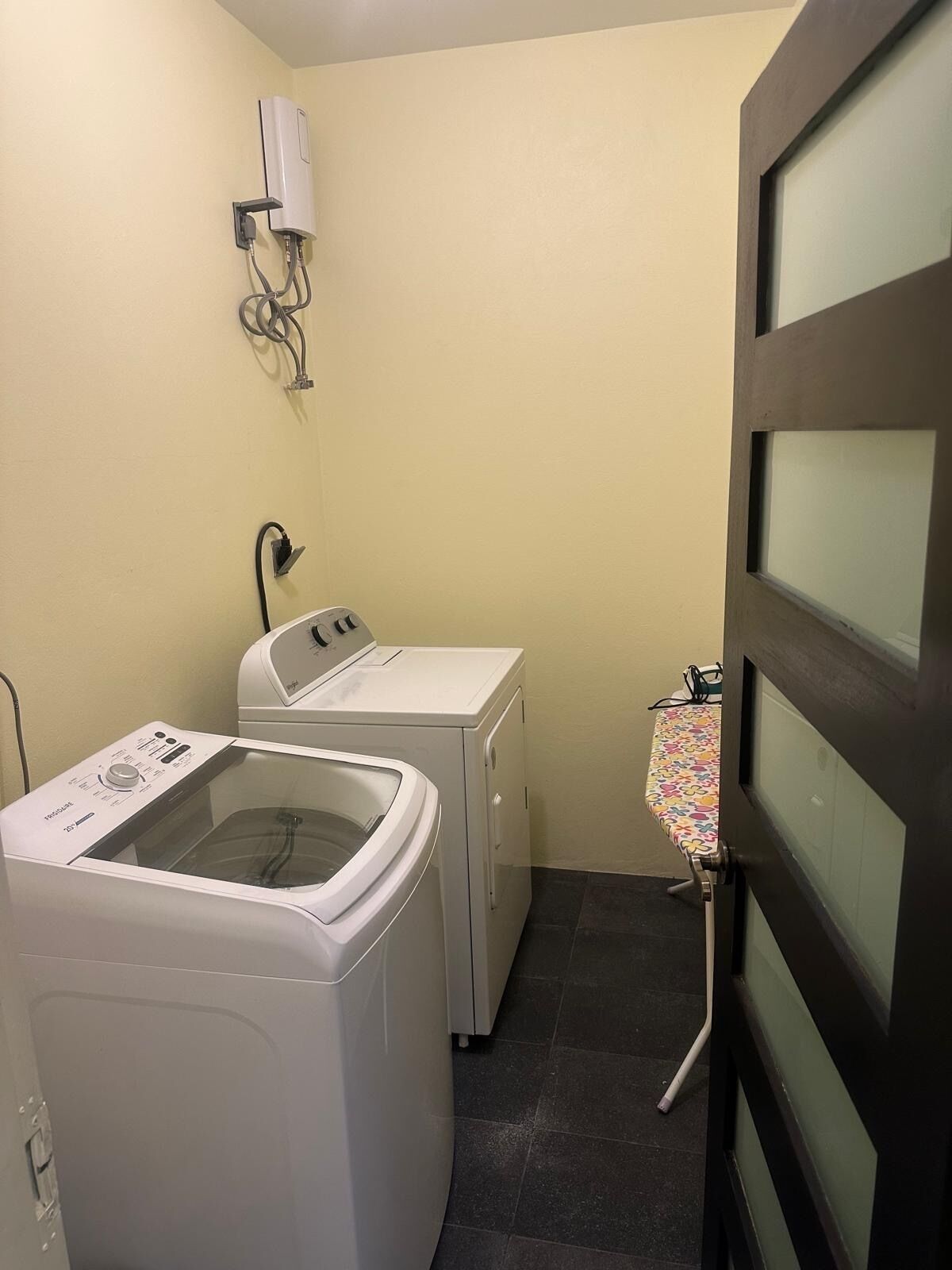 Un cuarto de lavandería con lavadora y secadora de carga frontal, una tabla de planchar pequeña con mantel floreado y un calentador fijado a la pared.