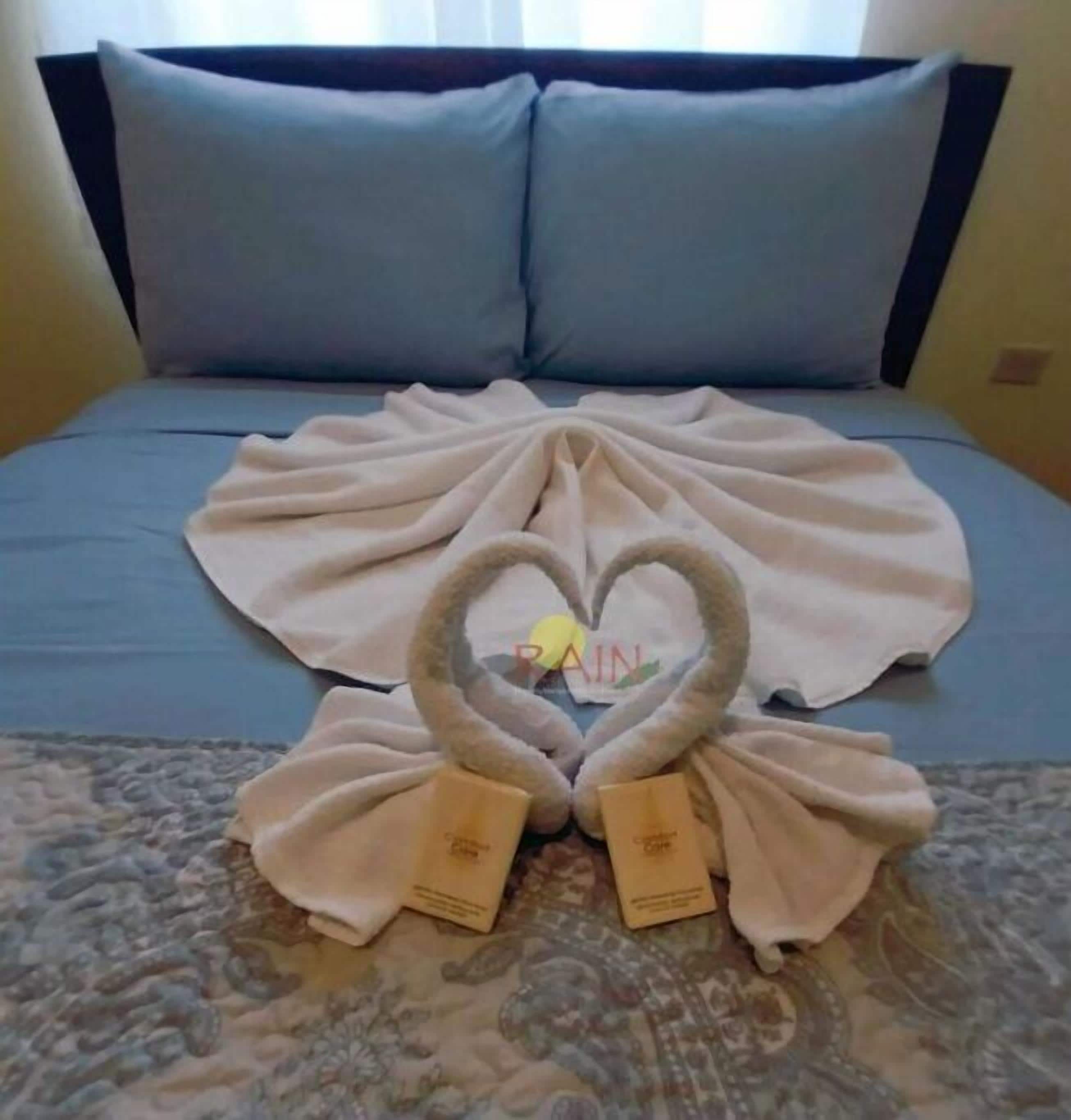 Un lit avec une couvre-lit bleue, deux oreillers et une décoration en forme de cœur, réalisée avec des serviettes et agrémentée d’étiquettes.