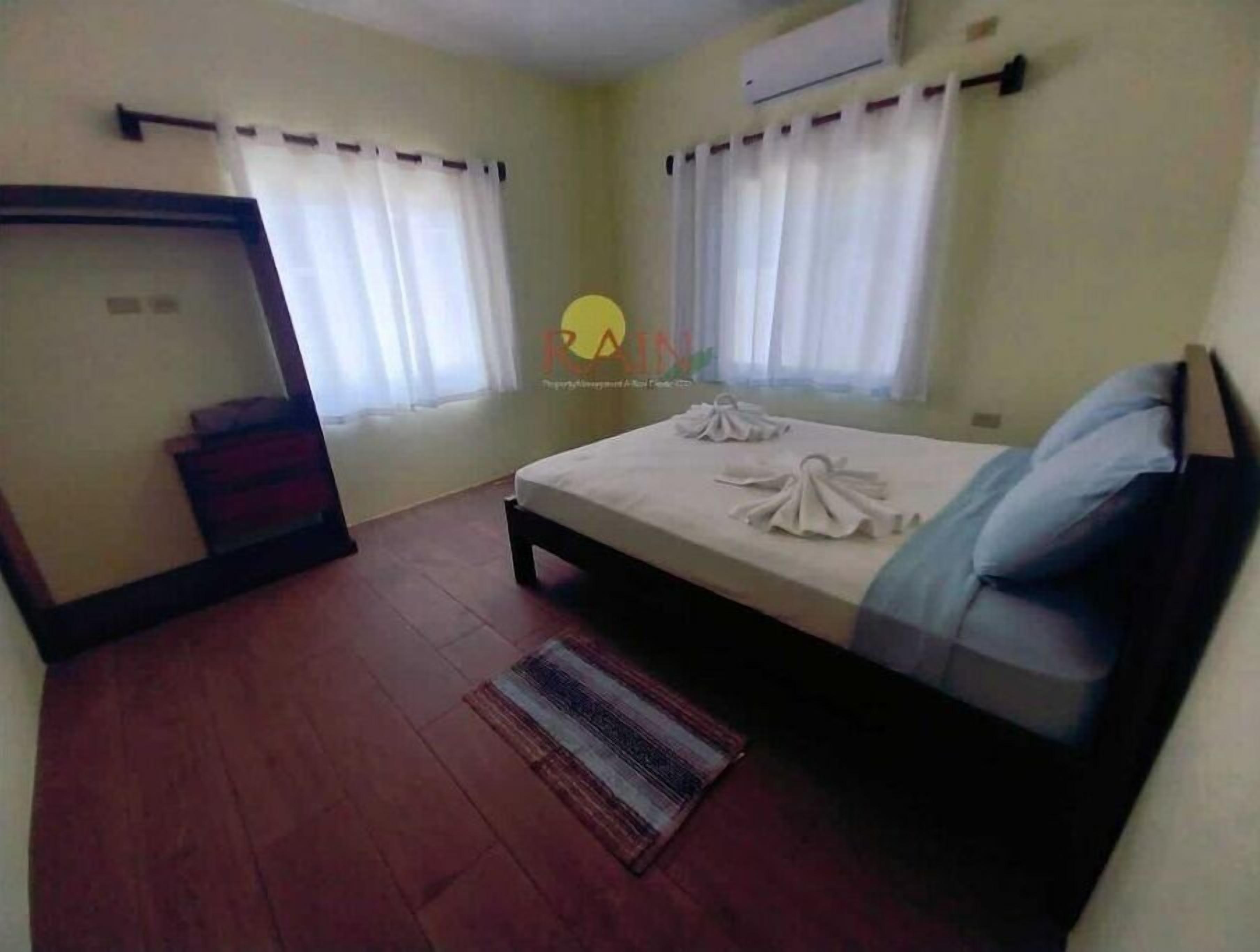 A bedroom with a bed, two windows with curtains, a wooden floor, and a small rug.