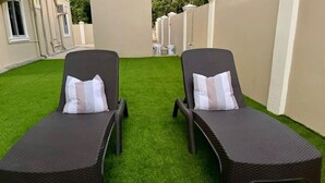 Deux fauteuils de jardin en osier foncé, dotés de coussins à rayures blanches, sont placés sur une pelouse verdoyante et bien entretenue, devant un mur beige orné d’une fenêtre et d’un appareil de climatisation.