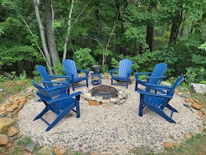 Un ensemble de chaises Adirondack bleues disposées en cercle autour d’un foyer central, le tout installé sur une surface de gravier agrémentée de rochers et de verdure.