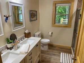 Une salle de bain avec un lavabo blanc, des toilettes, une fenêtre donnant sur un espace verdoyant et un plancher en bois.