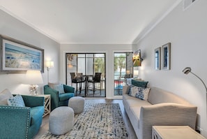 A modern living room with teal and beige furniture, a dining area with a glass door leading to a balcony, and artwork on the walls.