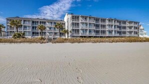 Un hotel frente al mar con varios pisos, ventanas amplias y una playa arenosa al frente.