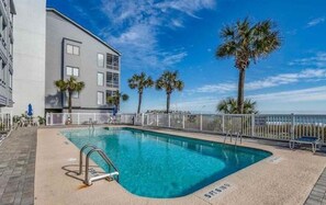 A swimming pool with a metal ladder, surrounded by a white fence, palm trees, and a building with balconies.