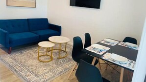 Un salón con un sofá azul, una mesa de centro, una mesa de comedor con sillas, una alfombra, un cuadro de una playa y un televisor en la pared.
