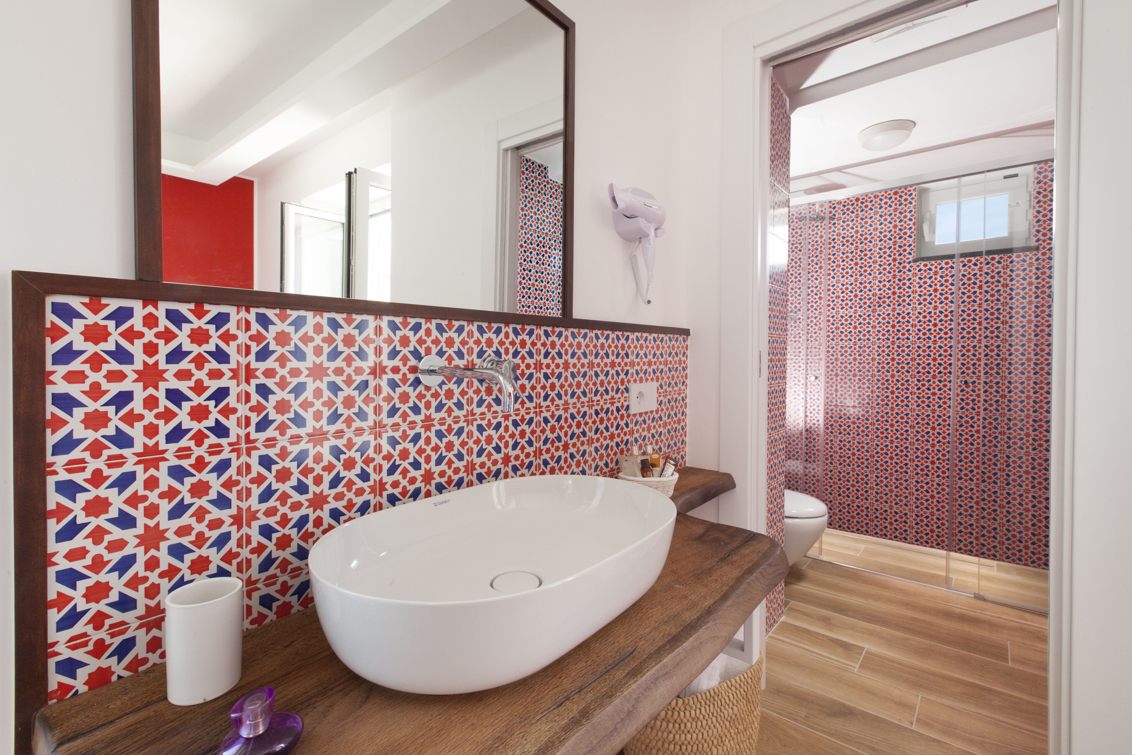 Un baño con un lavamanos grande de color blanco, encimera de madera, espejo y salpicadero de azulejos estampados.