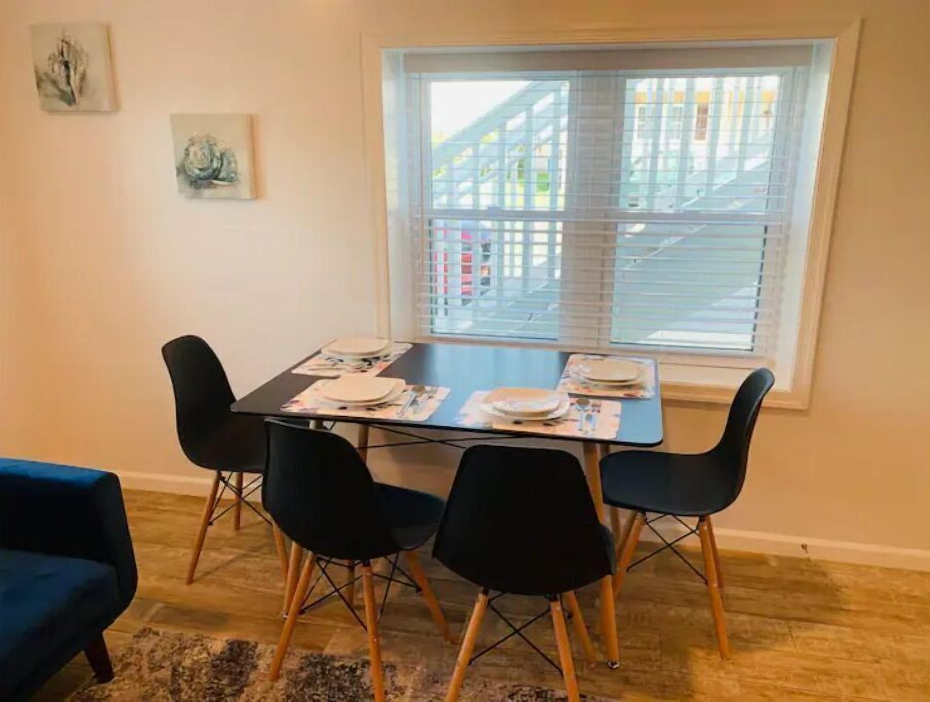 Un coin repas avec une table dressée pour quatre personnes, quatre chaises noires, un canapé bleu et une fenêtre avec des stores.