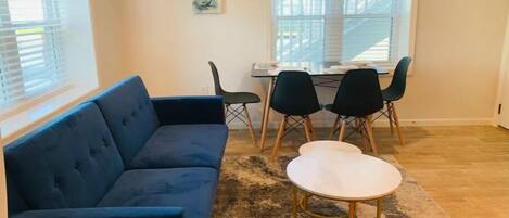 Ein Wohnzimmer mit einem blauen Sofa, einem weißen Couchtisch und einem Essbereich inklusive Tisch und Stühlen.