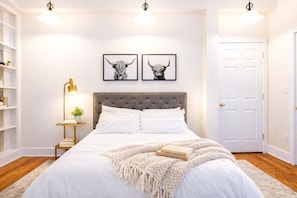Une chambre à coucher avec une tête de lit capitonnée, du linge de lit blanc, deux tableaux représentant des animaux encadrés au mur, une lampe de chevet et une porte.