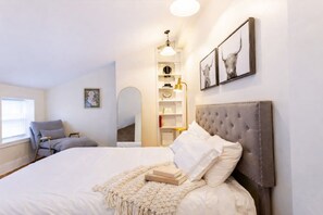 A bedroom with a large bed, a bedside table, a chair, a bookshelf, and a mirror.