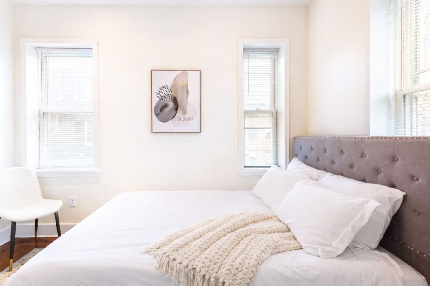 Un dormitorio con una cama grande, cabecera, una obra de arte enmarcada en la pared y dos ventanas con persianas.