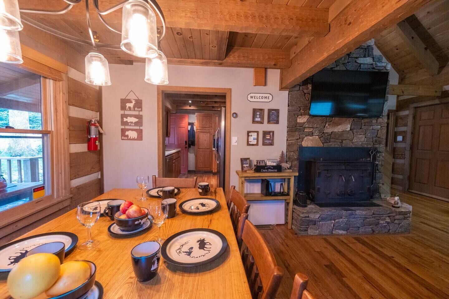 A wooden dining table set with plates, glasses, and fruit, a stone fireplace, and a 'WELCOME' sign in a cabin-style interior.
