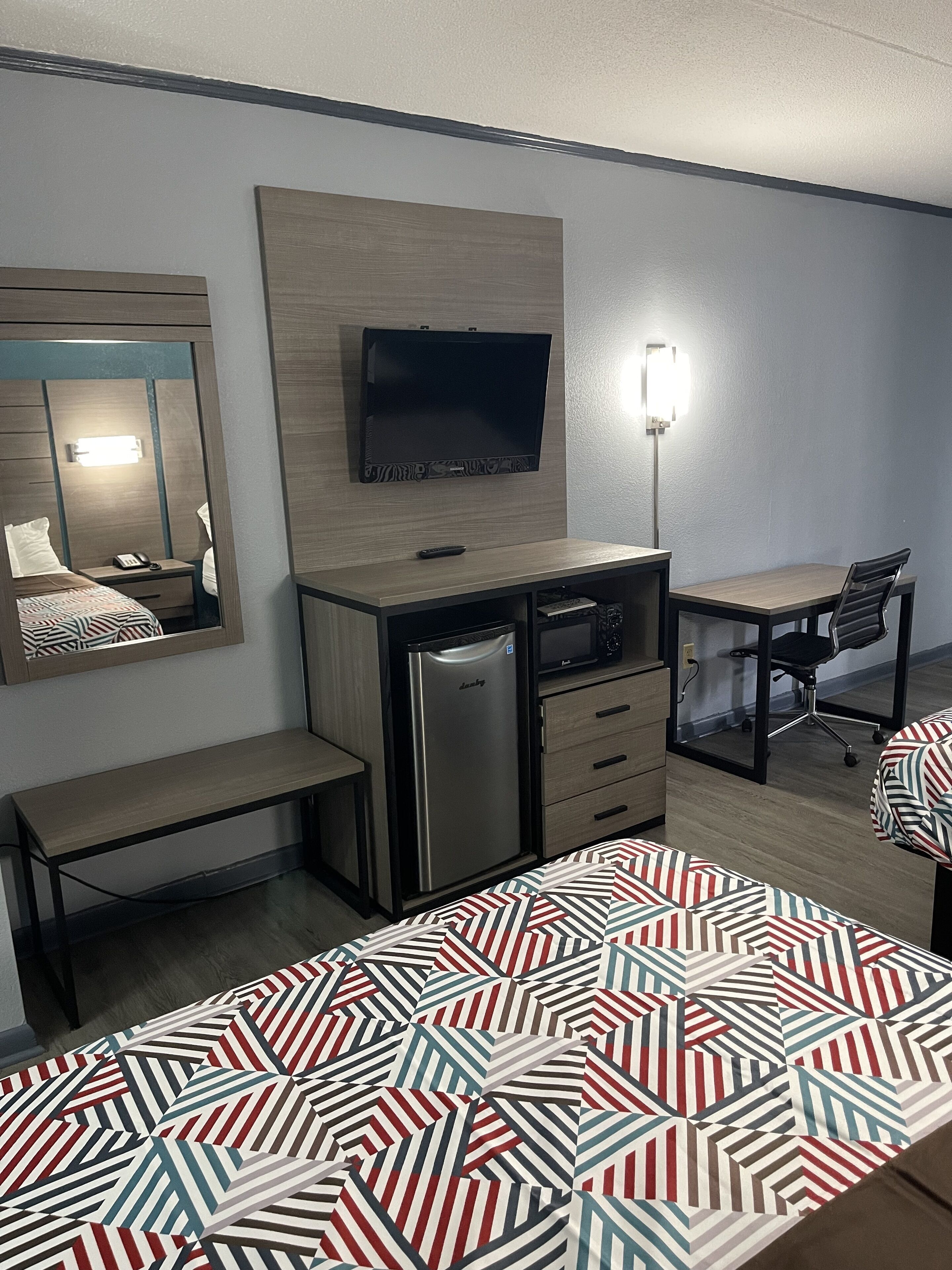 A compact hotel room with a bed, a desk, a refrigerator, a television, and a mirror.