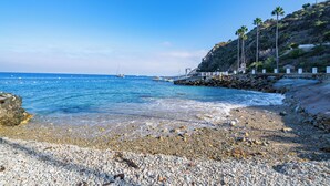 Ein Kiesstrand mit klarem, blauem Wasser, einer felsigen Küstenlinie und einer entfernten Bootsansicht.