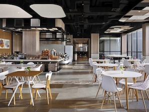 A modern dining area with multiple tables and chairs, a kitchen in the background, and large windows providing natural light. - Ibis Yogyakarta Airport Kulon (Temon)