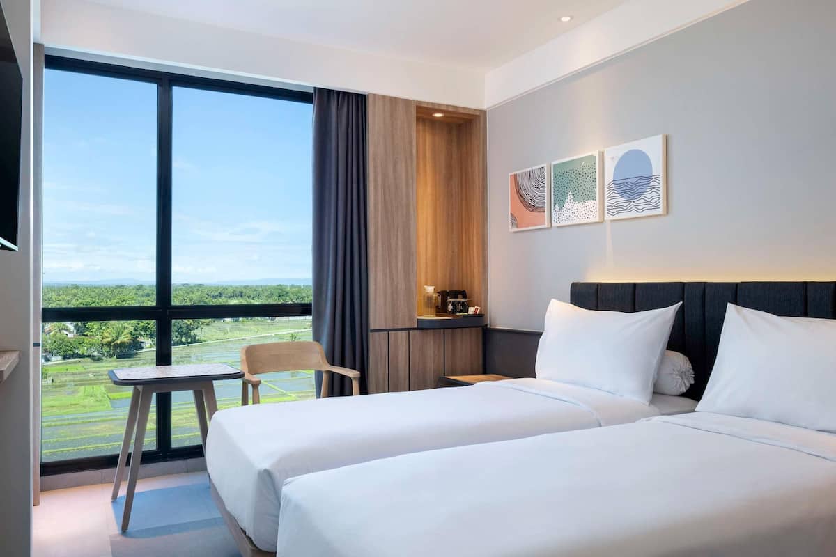 A hotel room with two beds, a small table, a chair, and a view of a green landscape through large windows.