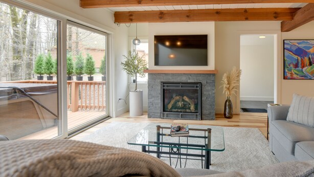 A living room with a fireplace, a television mounted above it, and a large window overlooking a deck with potted plants. - Private Hot Tub & Deck: North Conway Retreat (North Conway)