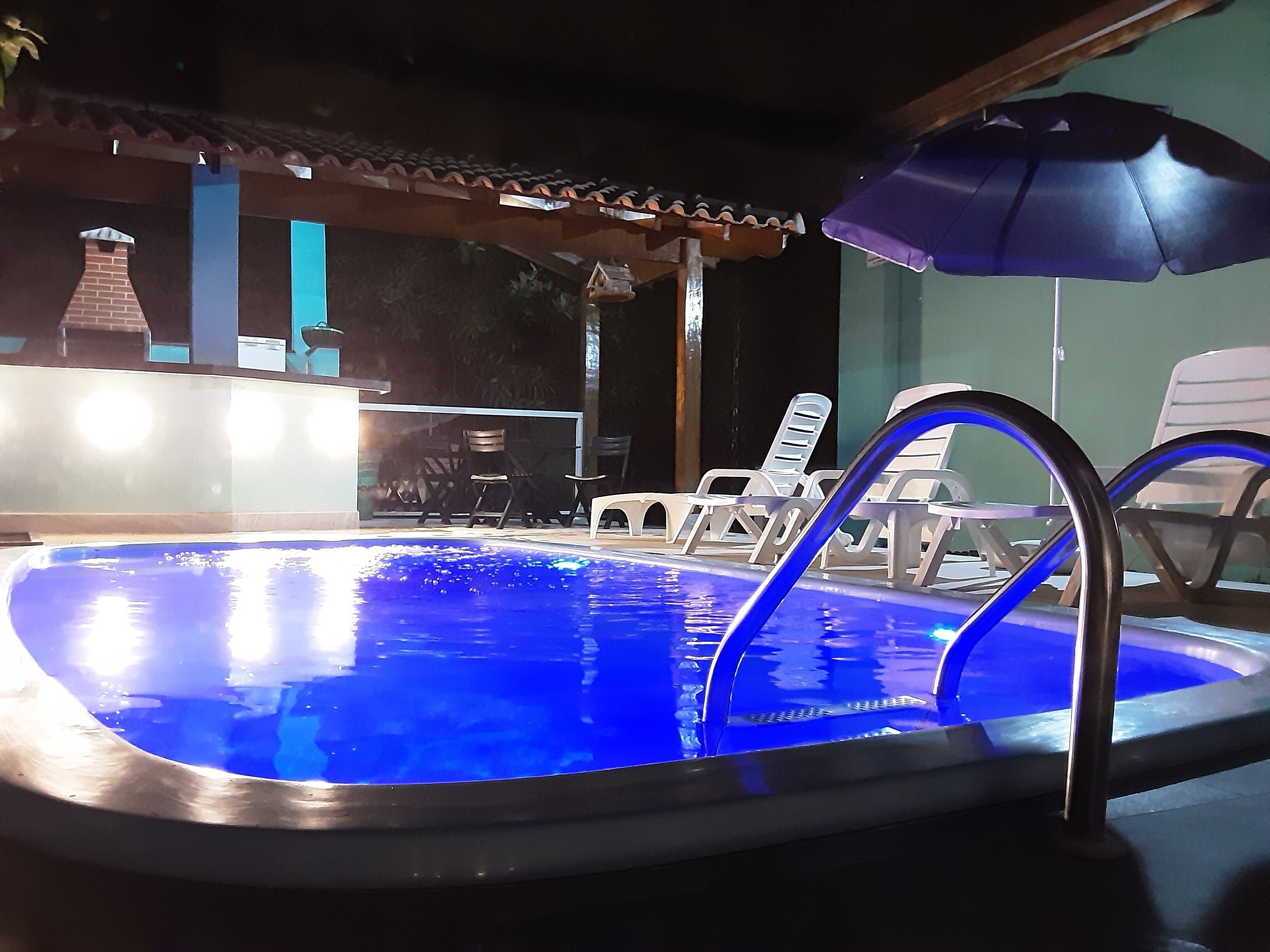 An outdoor swimming pool with a blue light, surrounded by lounge chairs and umbrellas.