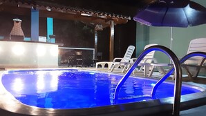 An outdoor swimming pool with a blue light, surrounded by lounge chairs and umbrellas.