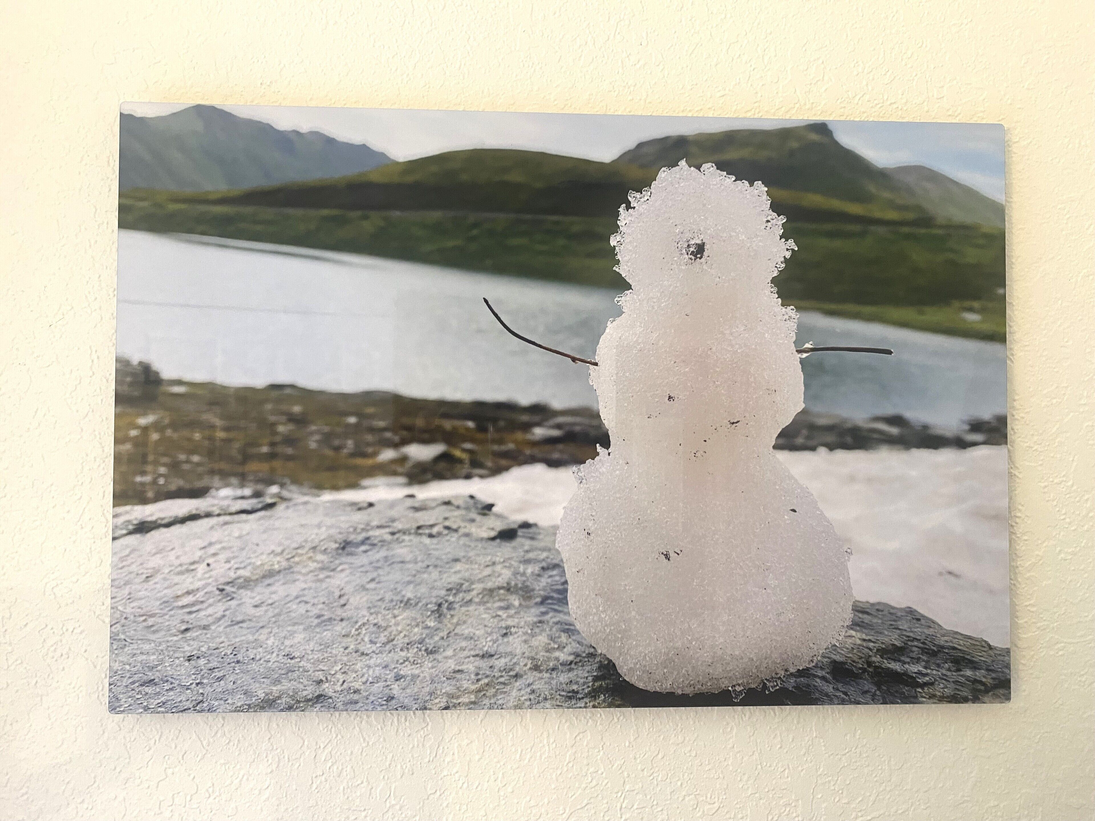 A snowman on a rock with a lake and mountains in the background.