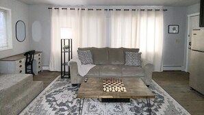 Un salon avec un canapé beige, une table basse sur laquelle sont posés des pièces d’échecs, un tapis à motifs, une table d’appoint avec une lampe et un réfrigérateur.