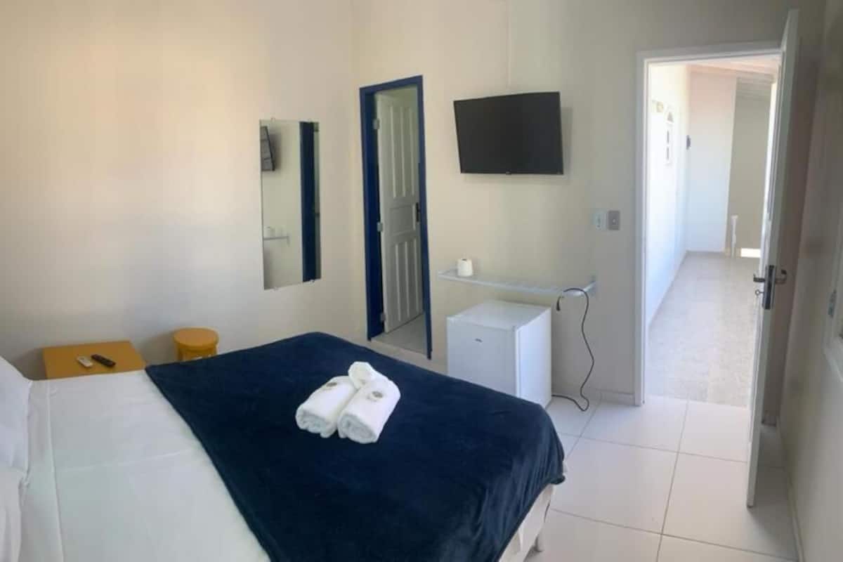 Et hotelværelse med en seng, et natbord, et spejl, et fjernsyn, et badeværelse med vask og toilet, samt en dør til et andet rum.