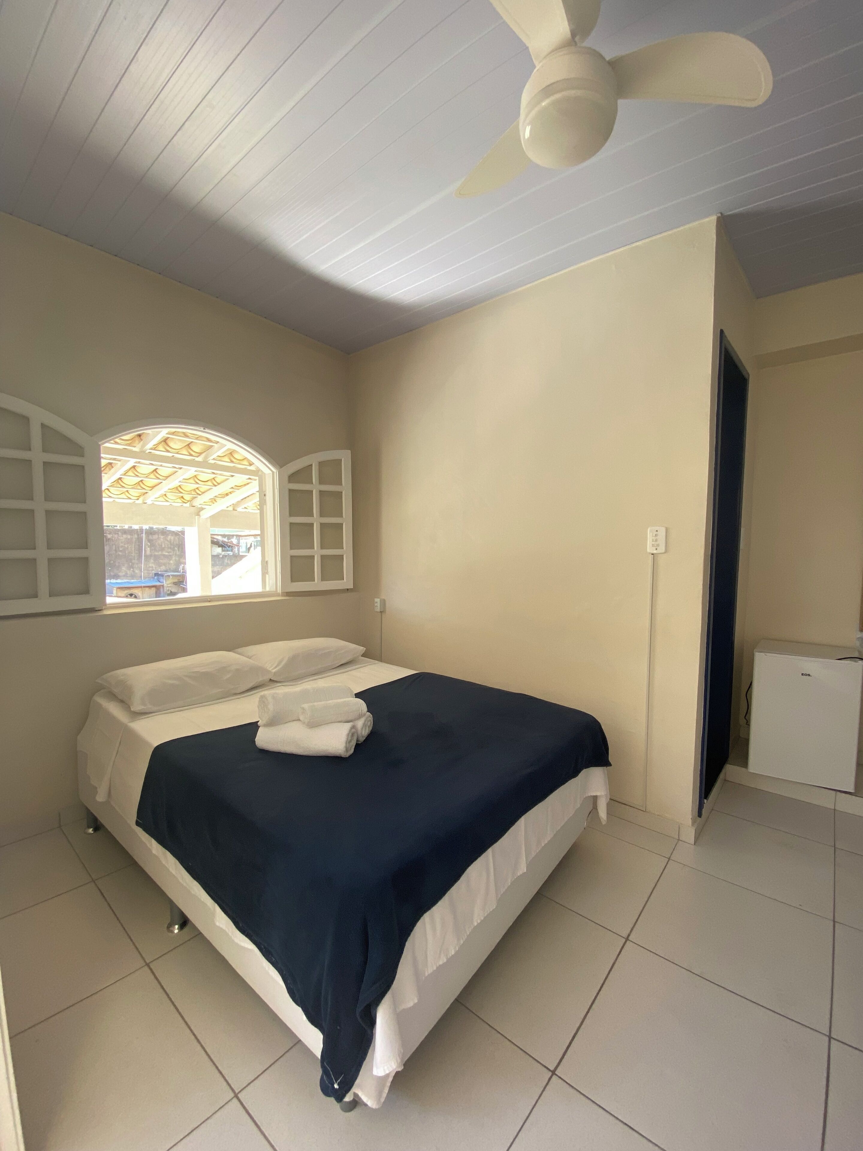 Une chambre avec un lit, un ventilateur de plafond, une fenêtre avec des volets et un petit réfrigérateur.