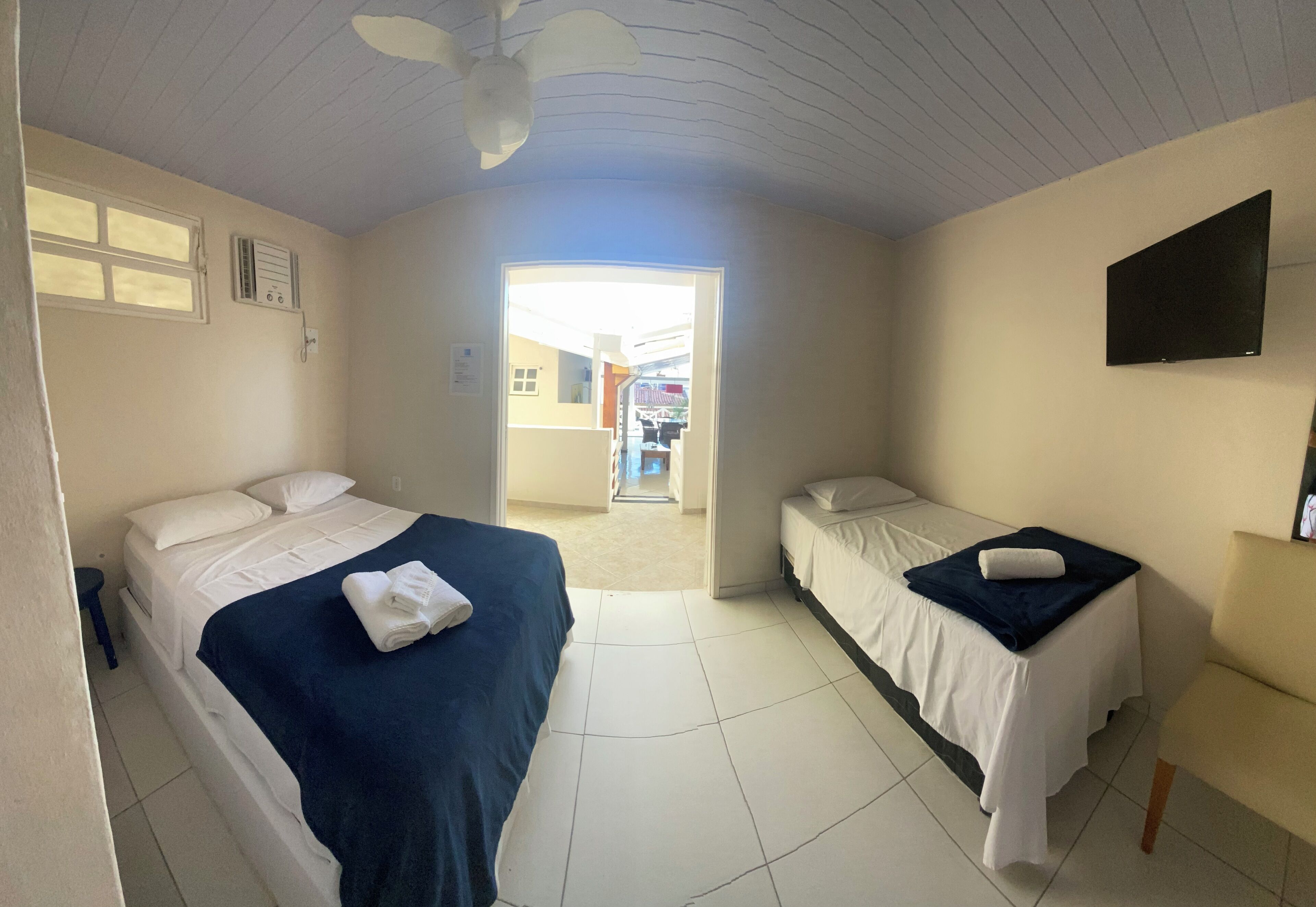 Ein Hotelzimmer mit zwei Betten, einem Deckenventilator, einem Flachbildfernseher und Blick durch eine offene Tür nach draußen.
