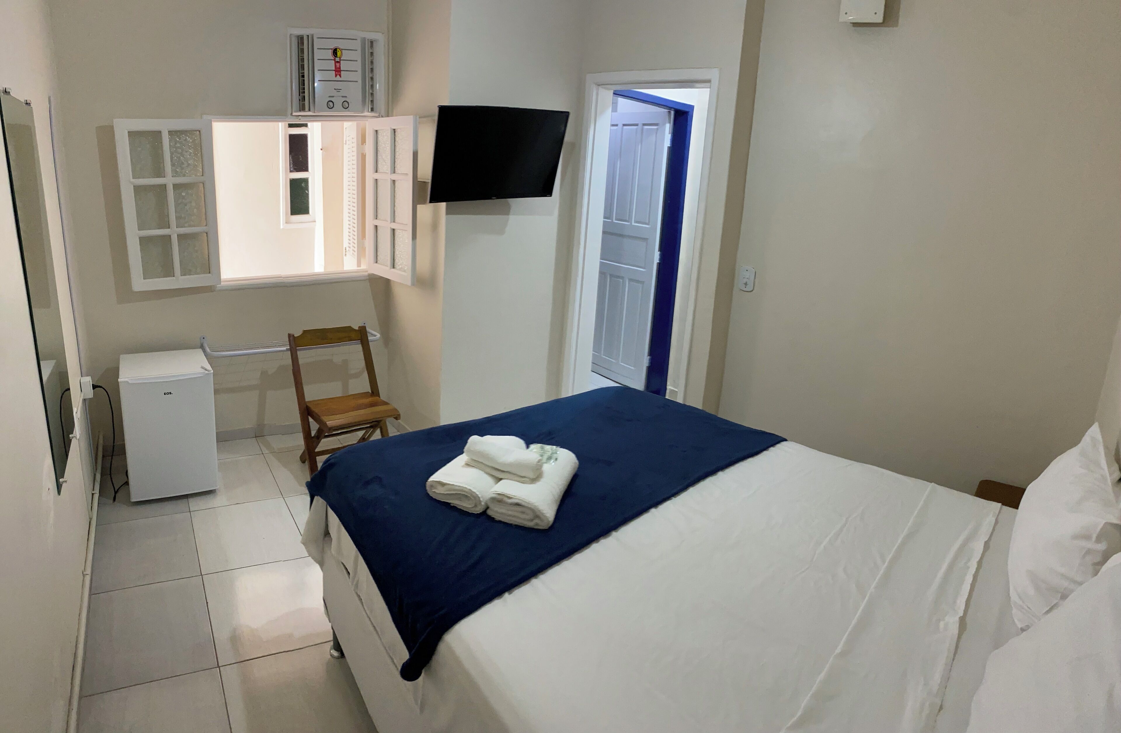 Un dormitorio con cama, un refrigerador blanco pequeño, un televisor montado en la pared y una ventana con vistas al exterior.